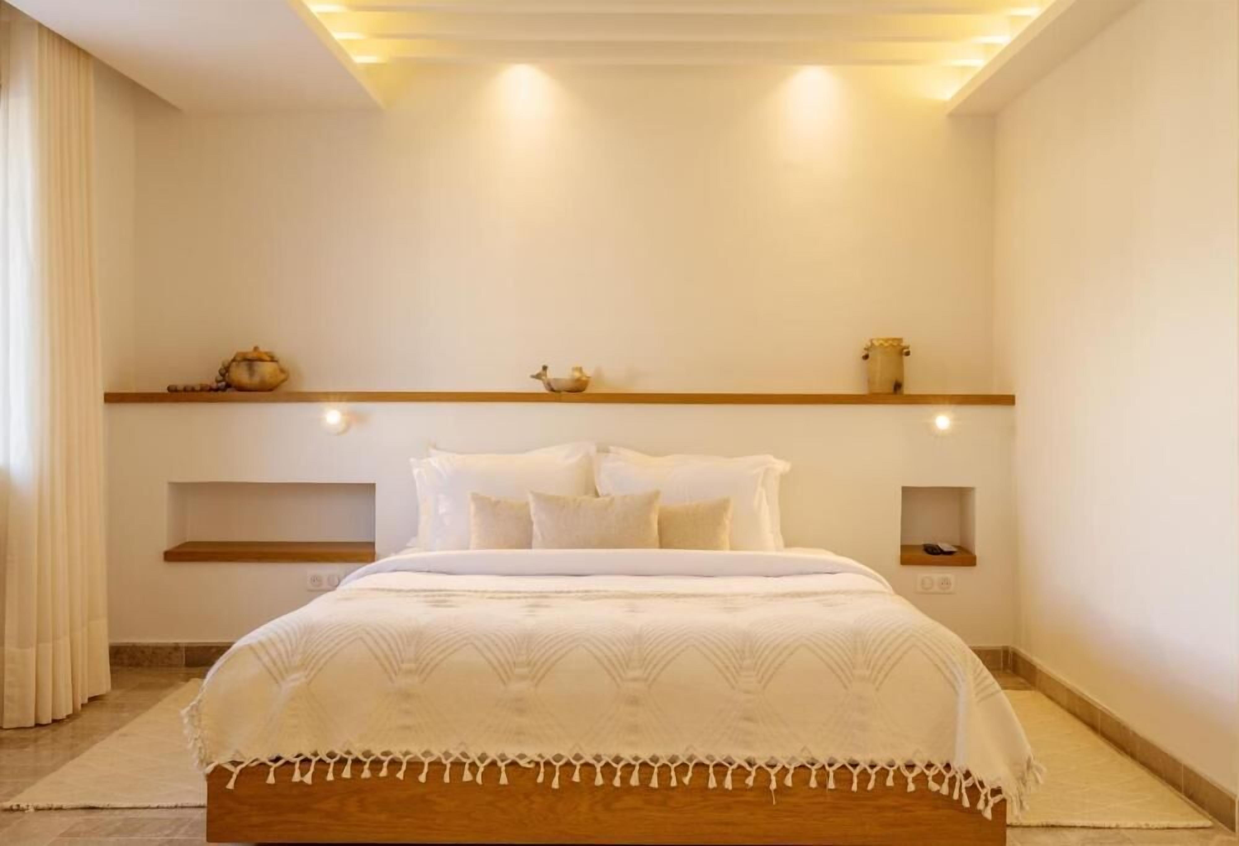 Un dormitorio con estructura de cama de madera, ropa de cama blanca y estantes empotrados con objetos decorativos.