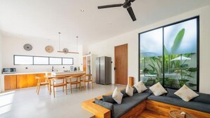 A modern living room with a wooden sectional sofa, a dining area with a wooden table and chairs, and a large window with a view of greenery. - Casa Riva by GORO (Bali)
