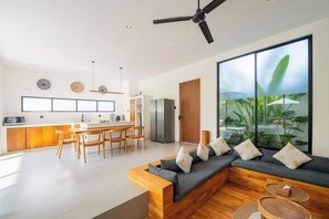 Ein modernes Wohnzimmer mit einem modularen Sofa aus Holz, einem Essbereich mit Holztisch und -stühlen sowie einem großen Fenster mit Blick auf Grünflächen.