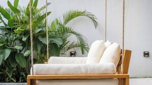 A wooden swing chair with white cushions against a white wall, surrounded by green plants. - Casa Riva by GORO (Bali)