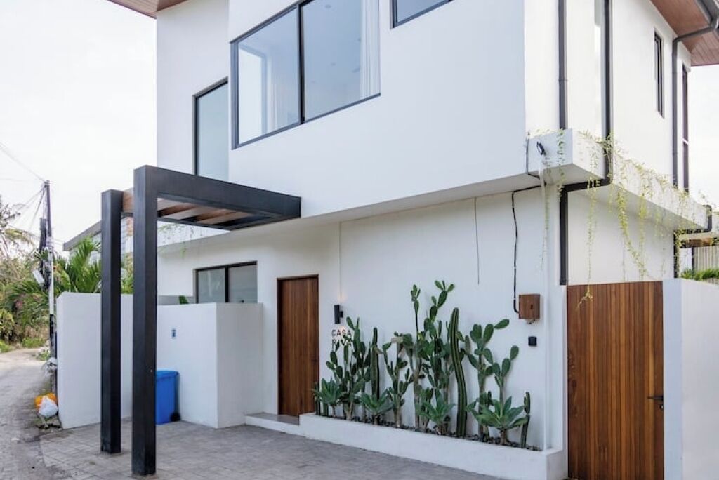 Ein modernes, zweistöckiges Haus mit weißer Fassade, großen Fenstern und einer Holztür. Entlang der Vorderwand sind Kakteen gepflanzt.
