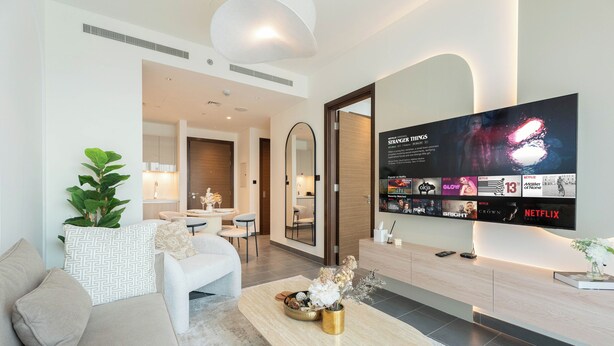A modern living room with a flat-screen TV displaying a movie poster, a sofa, a coffee table with decorative items, and a minimalist design. - Chic 1BR Apartment - The Crest Dubai (Dubai)