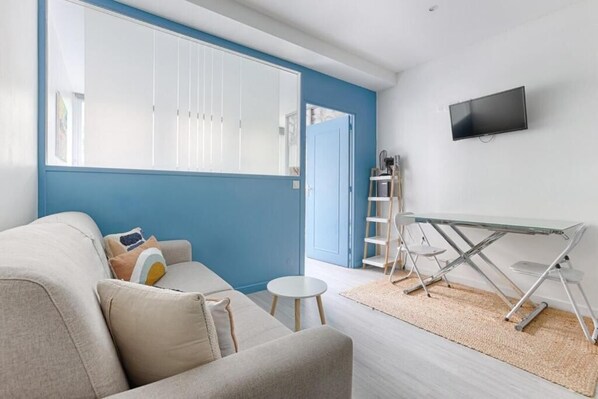 A modern living room with a sofa, a small round table, a glass-top coffee table, a TV mounted on the wall, and a blue accent wall. - Charming apartment Gambetta - Saint Fargeau II (Paris)
