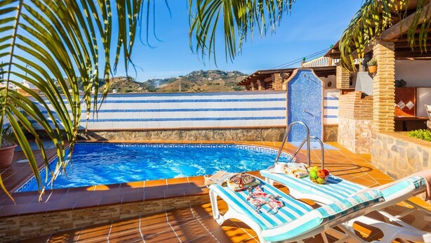 En swimmingpool med flisepyntet solterrasse, liggestole og palmer.