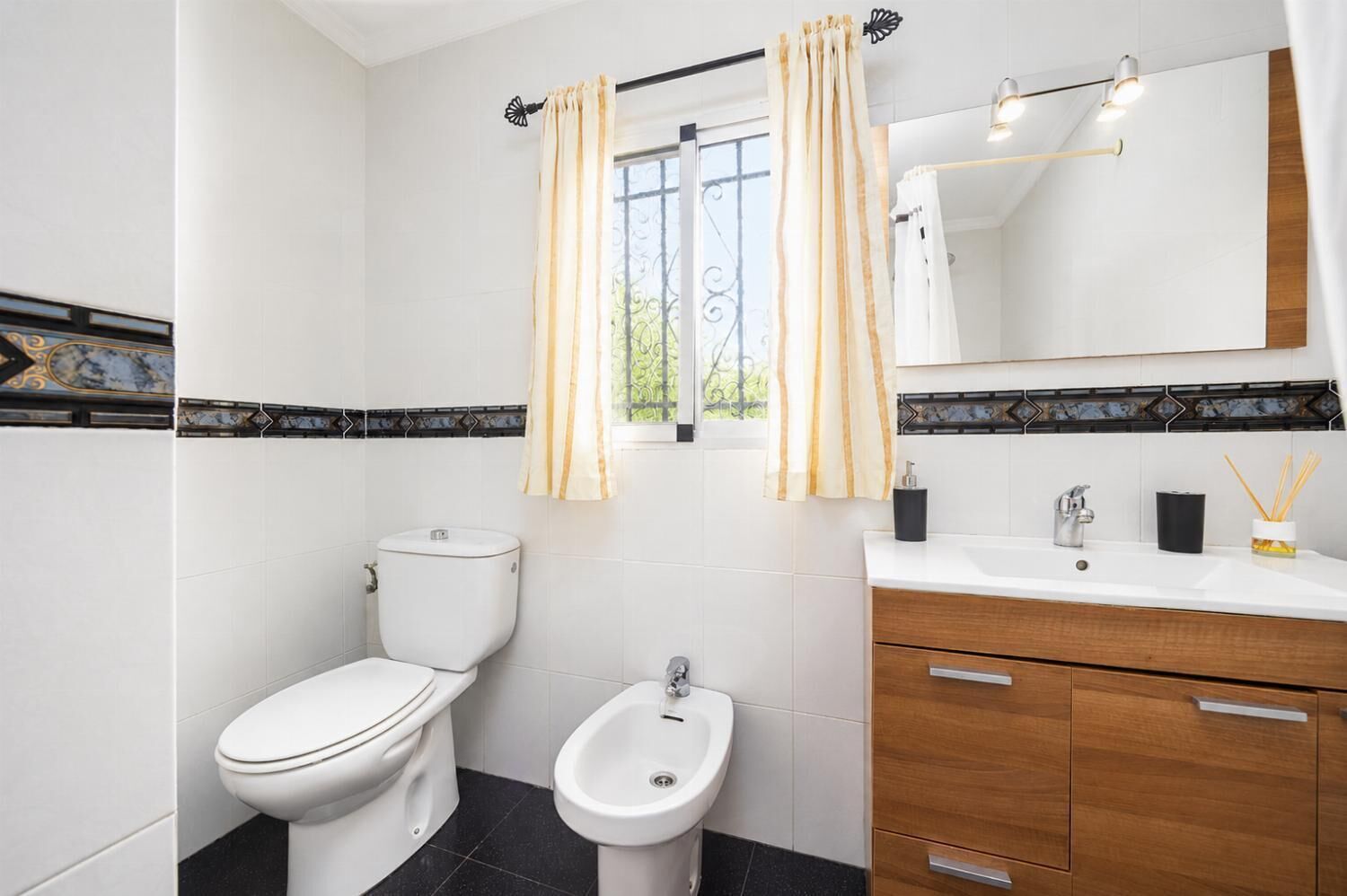 A bathroom with a toilet, bidet, vanity with a mirror, and a window with curtains.