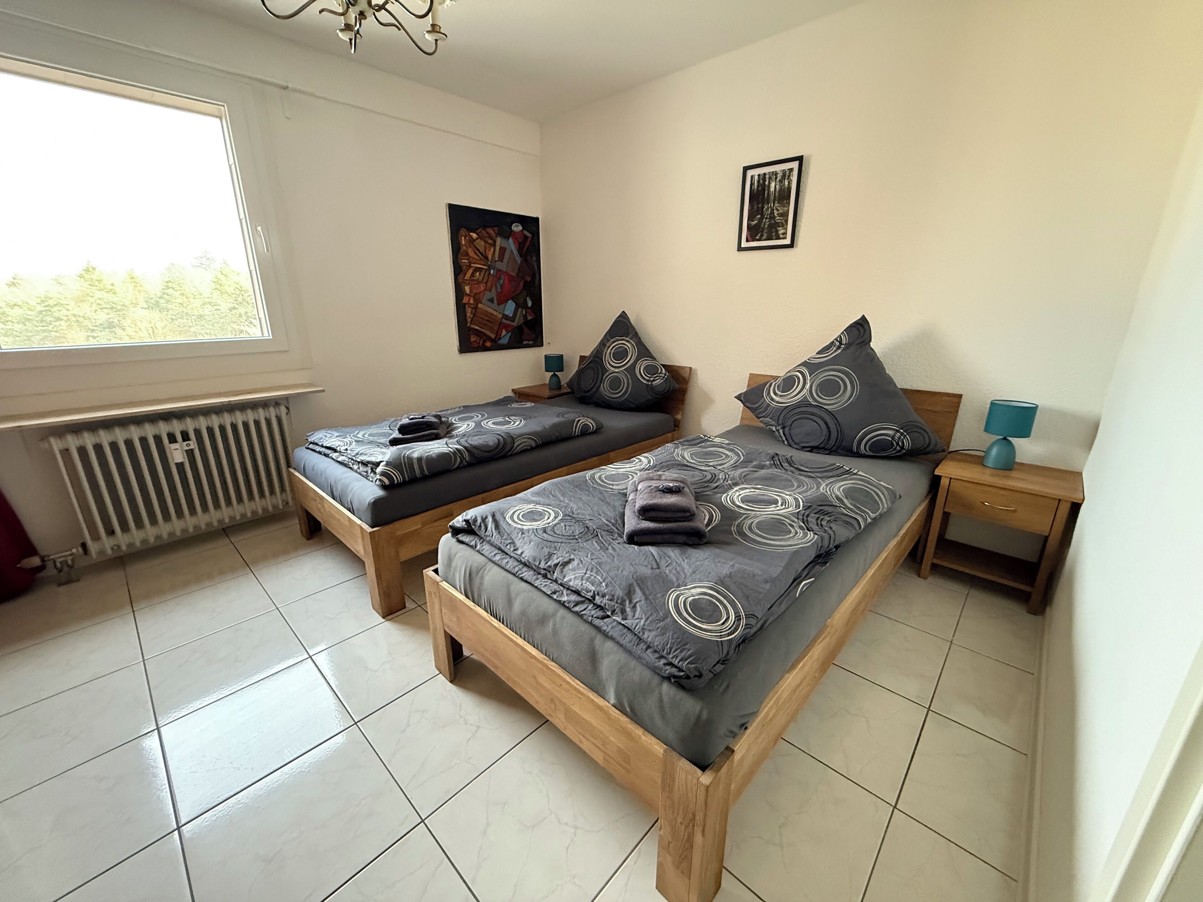 Un dormitorio con dos camas de madera, un radiador, una ventana y una mesita de noche con una lámpara.