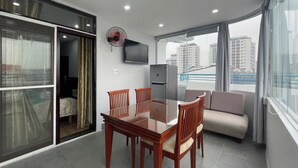 A modern living room with a glass dining table, wooden chairs, a sofa, a TV, and a balcony with a city view.
