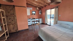Een slaapkamer met een bed, een houten ladder, een kitchenette met fornuis en een raam met gordijnen.