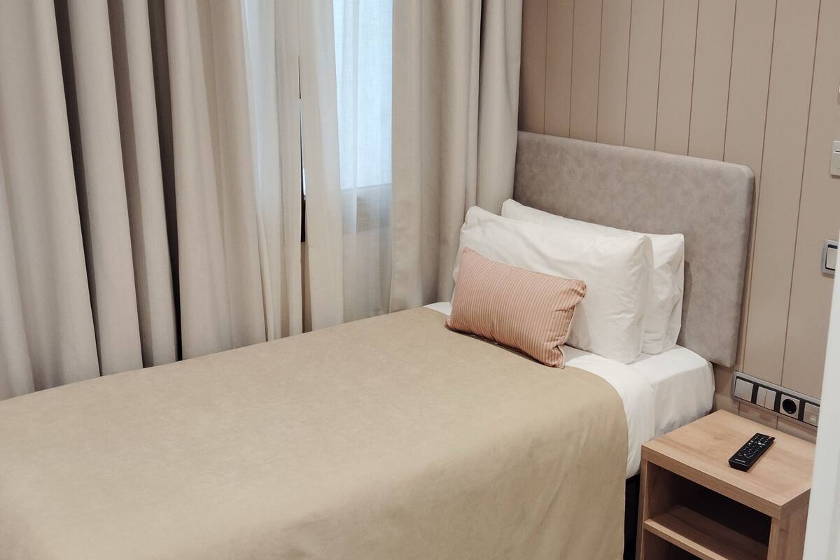 A neatly made bed with a beige bedspread, a single white pillow, and a pink pillow. There is a wooden nightstand with a remote control on it.