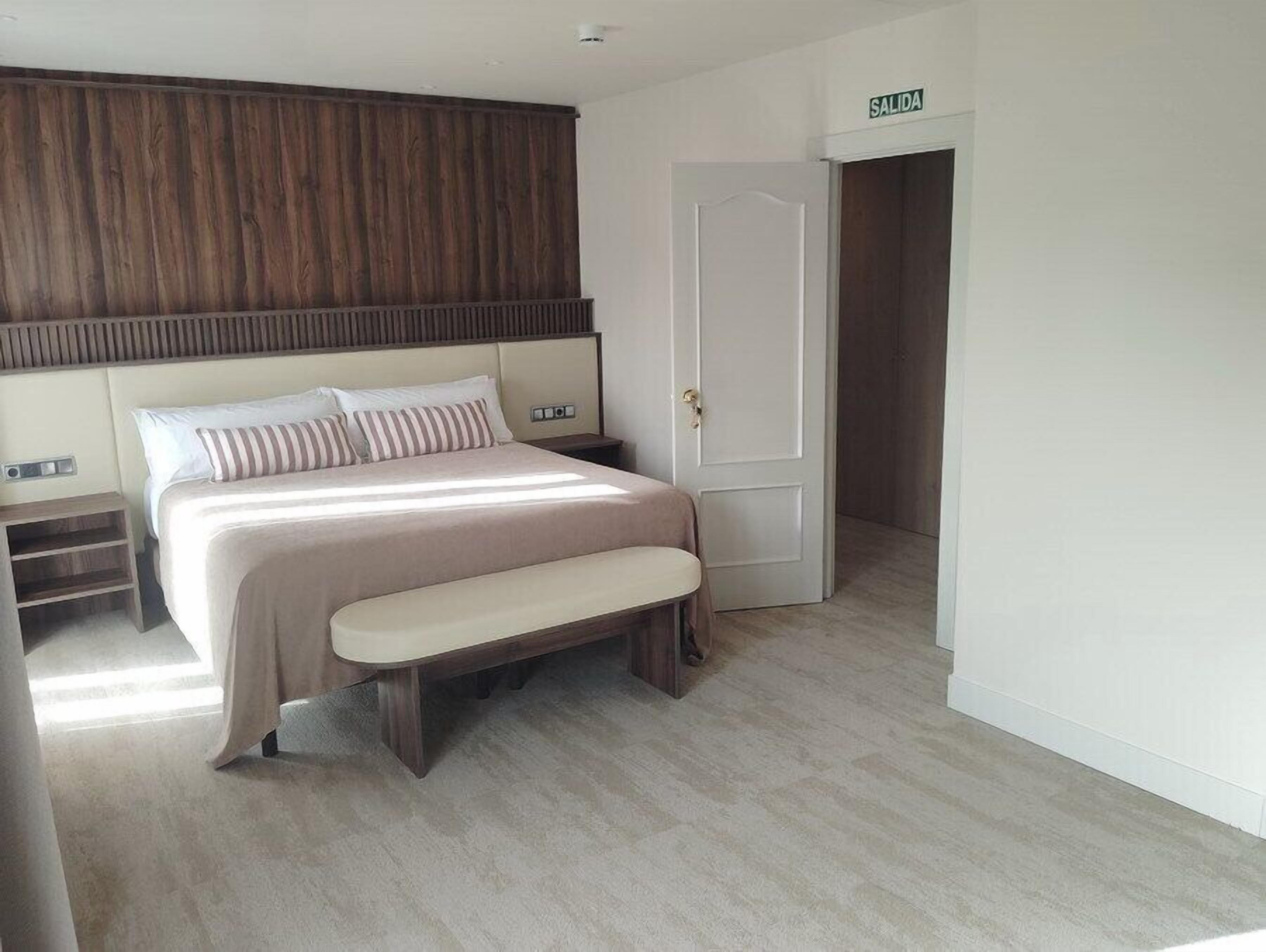 A bedroom with a bed, bench, bedside table, and a door labeled 'SALIDA'.