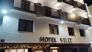 Ett hotell som heter HOTEL VÉLEZ, med en modern fasad, flera balkonger och en skylt som visar var parkeringsplatsen finns.