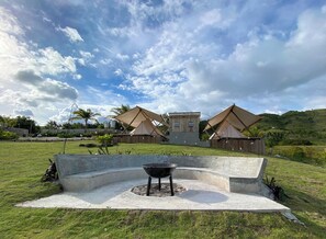 A grassy area with a concrete barbecue pit, surrounded by two wooden structures with pitched roofs, likely tents or cabins, set against a backdrop of hills and a partly cloudy sky. - Glamping at Kalumo Lodge - Tent 2 (Oslob)
