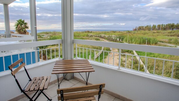 A balcony with a wooden table and two chairs, overlooking a grassy landscape and the sea. - Sea Apartment - with shared Pool (Villaggio del Golfo)