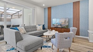 Un salon moderne avec un canapé gris, des fauteuils à motifs, une table basse et un téléviseur à écran plat fixé sur un mur à texture.