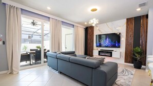 Una sala de estar moderna con un sofá gris, un televisor de pantalla plana y un comedor visible a través de puertas de cristal.