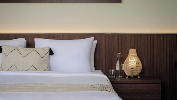 Una cama bien tendida con sábanas blancas y dos almohadas, una mesita de noche con lámpara y un panel decorativo en la pared.