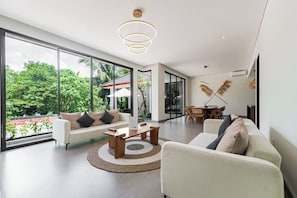 A spacious living room with a large sliding glass door leading to a balcony, a white sofa, a coffee table, and a dining area with a table and chairs. - Villa Jenggala by GORO (Kabupaten Gianyar)