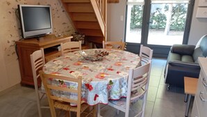 A dining area with a round table covered by a floral tablecloth, surrounded by wooden chairs. A television is mounted on the wall above the table.