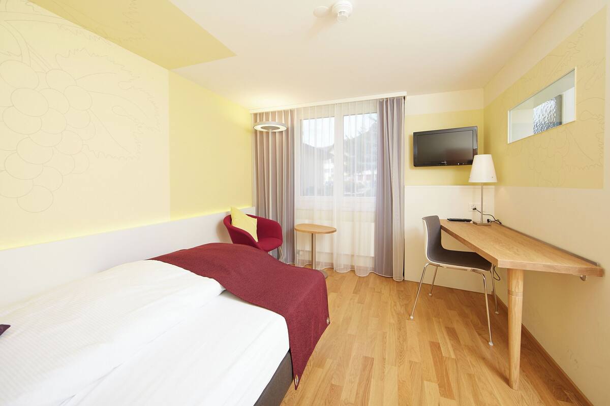 A hotel room with a bed, a desk, a chair, a TV, and a window with curtains.