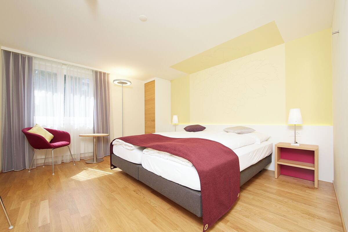 A modern hotel room with a bed, a red chair, a bedside table with a lamp, and a large window with curtains.