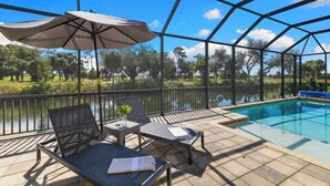 An outdoor pool area with lounge chairs and an umbrella, surrounded by a metal fence and a view of a lake and trees. - Seaside Villa Golfers Delight South facing, quiet location, privacy, heated pool (Cape Coral)