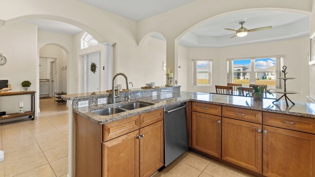 A spacious kitchen with granite countertops, wooden cabinets, and a dishwasher. There is a dining area with a table and chairs visible through an open archway. - Seaside Villa Golfers Delight South facing, quiet location, privacy, heated pool (Cape Coral)