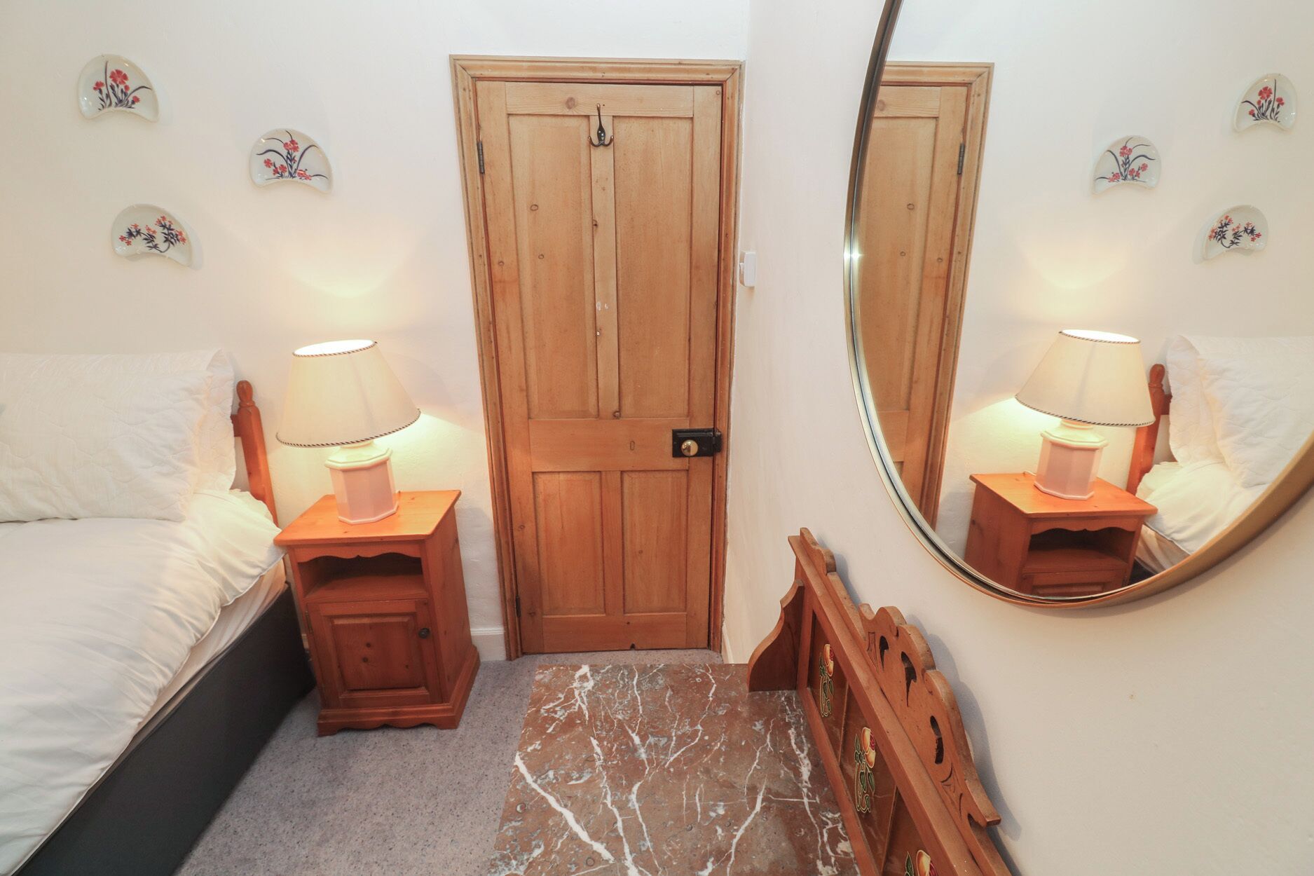 Ein Schlafzimmer mit Holztür, zwei Nachttischen inklusive Lampen, einem Bett mit weißer Bettwäsche, einem Teppich mit Marmoroptik sowie Wanddekorationen.