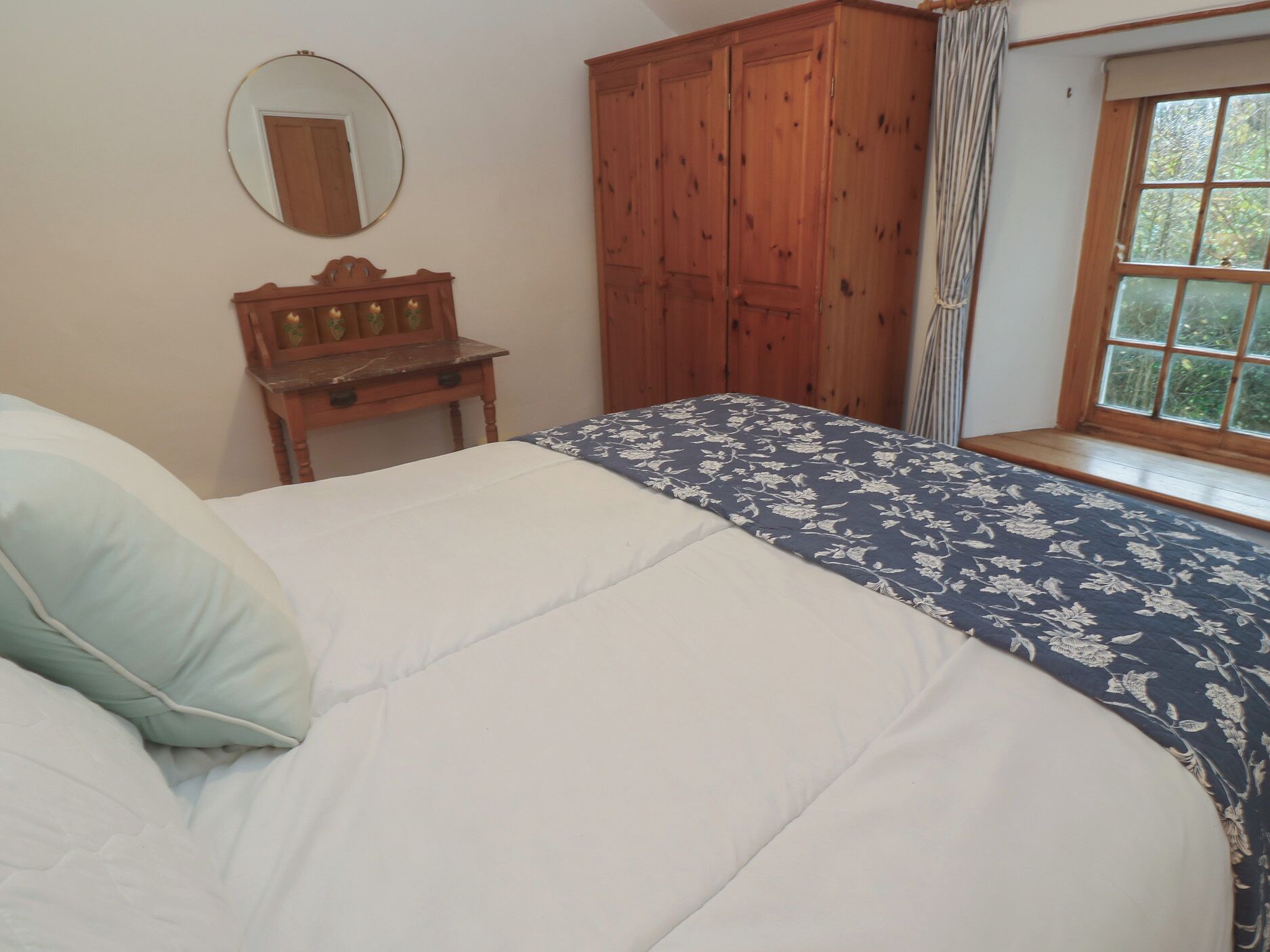 A bedroom with a bed, a wooden wardrobe, a small table with a mirror, and a window with curtains.