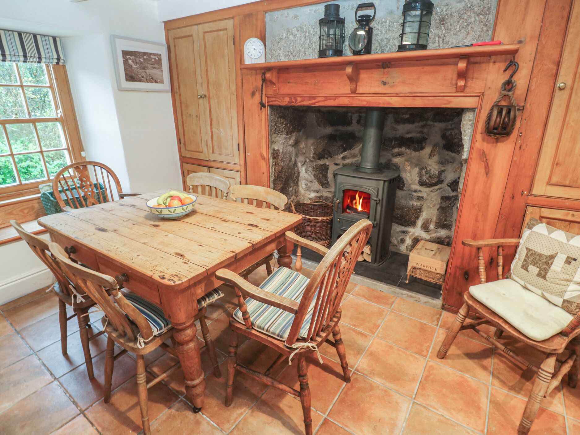 A rustic wooden dining area with a stone fireplace, wooden chairs, and a table set with a bowl of fruit.