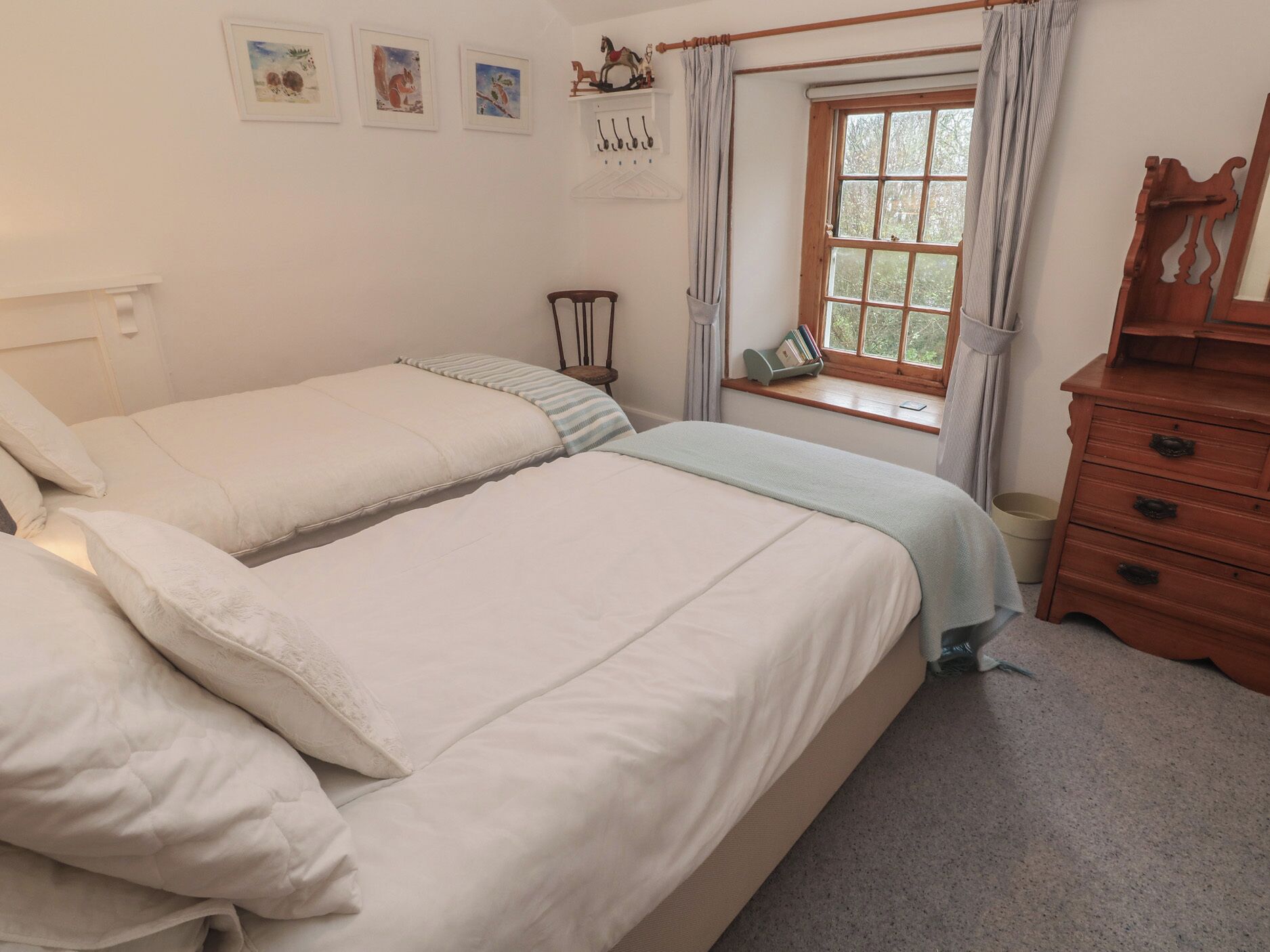 Une chambre avec deux lits, une commode en bois, une fenêtre avec des rideaux et des œuvres d’art accrochées au mur.