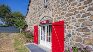 A stone building with a red shuttered entrance, a doormat, and a flower bed with pink flowers. - “Chambre Raphaël” Guest Room with Mountain View, Wi-Fi, and Air Conditioning (Singles)