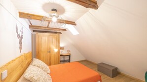 A bedroom with a sloped ceiling, a bed with an orange bedspread, a wooden wardrobe, a small table with a lamp, and a green chair. - “Chambre Raphaël” Guest Room with Mountain View, Wi-Fi, and Air Conditioning (Singles)