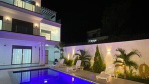 A modern two-story house with a swimming pool, outdoor seating, and a well-lit exterior. - Teranga Blue (Ngaparou)