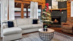 Un salon chaleureux avec un canapé, un fauteuil et une cheminée. Un sapin de Noël est décoré d’ornements et de guirlandes lumineuses.