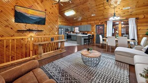 Un vaste espace de vie doté d’un plafond en bois, d’un téléviseur fixé au mur, d’un coin repas avec une table et des chaises, et d’une cuisine située à l’arrière-plan.