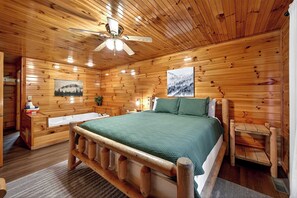 Quarto de cabana de madeira com uma cama, um banco de madeira, uma mesa pequena e um ventilador de teto.