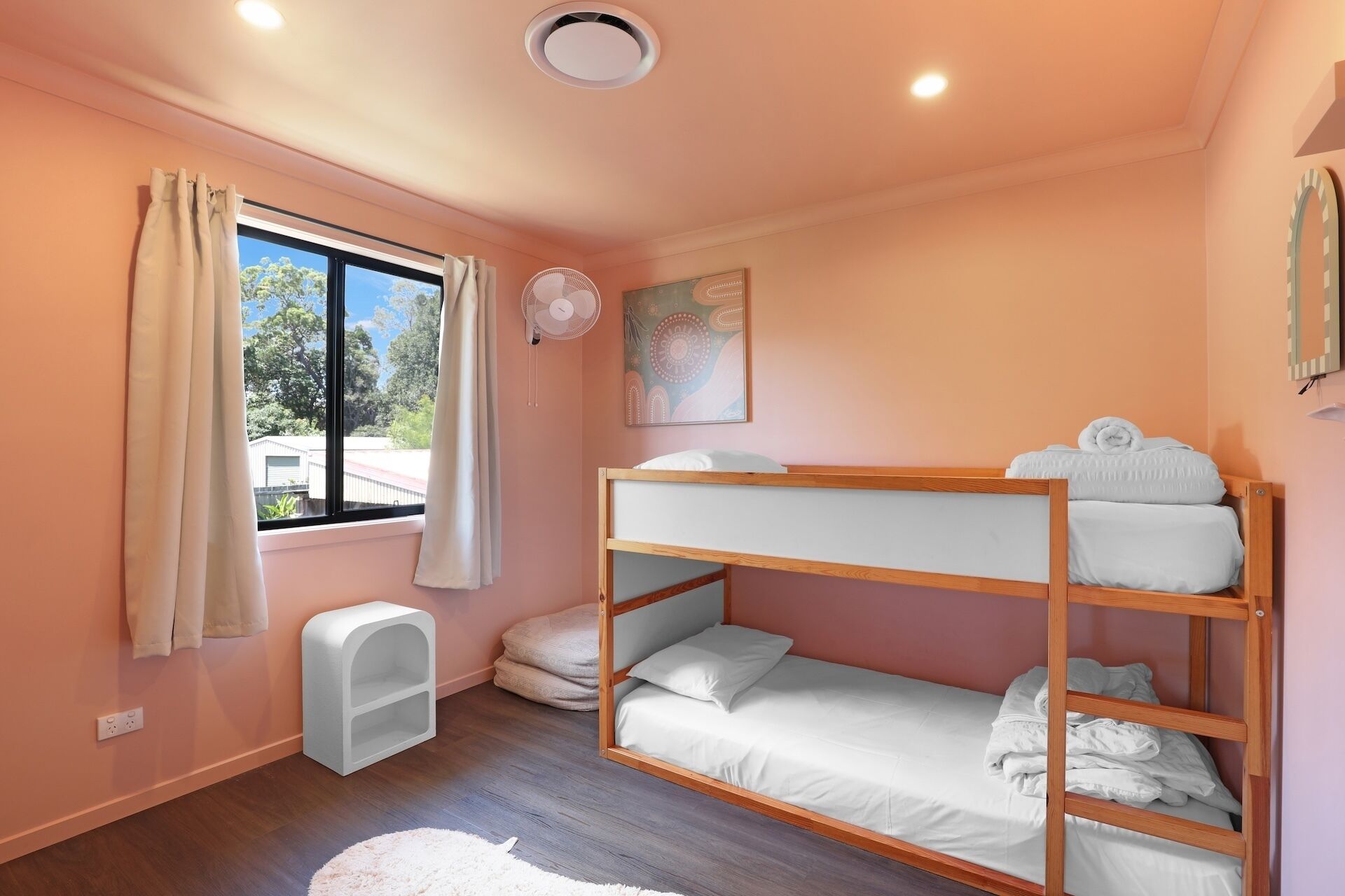 Ein Schlafraum mit Etagenbetten, einem Fenster, einem kleinen weißen Regal und einem Deckenventilator.