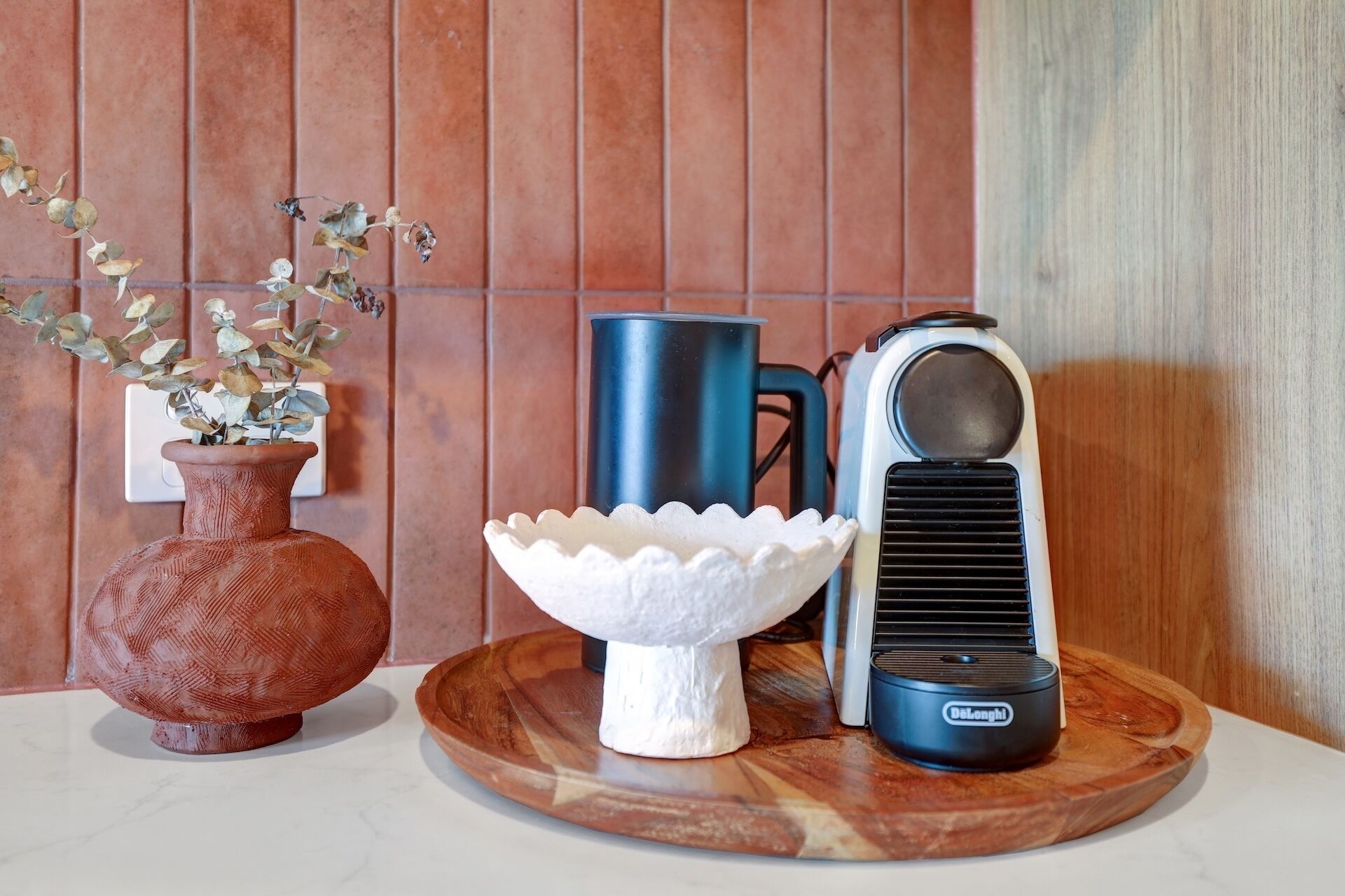 A rustic kitchen setting with a terracotta vase, a white ceramic bowl, a blue kettle, and a DeLonghi electric kettle on a wooden tray.