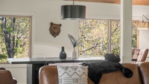 Un salon avec un canapé en cuir marron, un chat noir, un coussin à motifs, un vase avec une plante et une décoration murale représentant une tête d’ours.