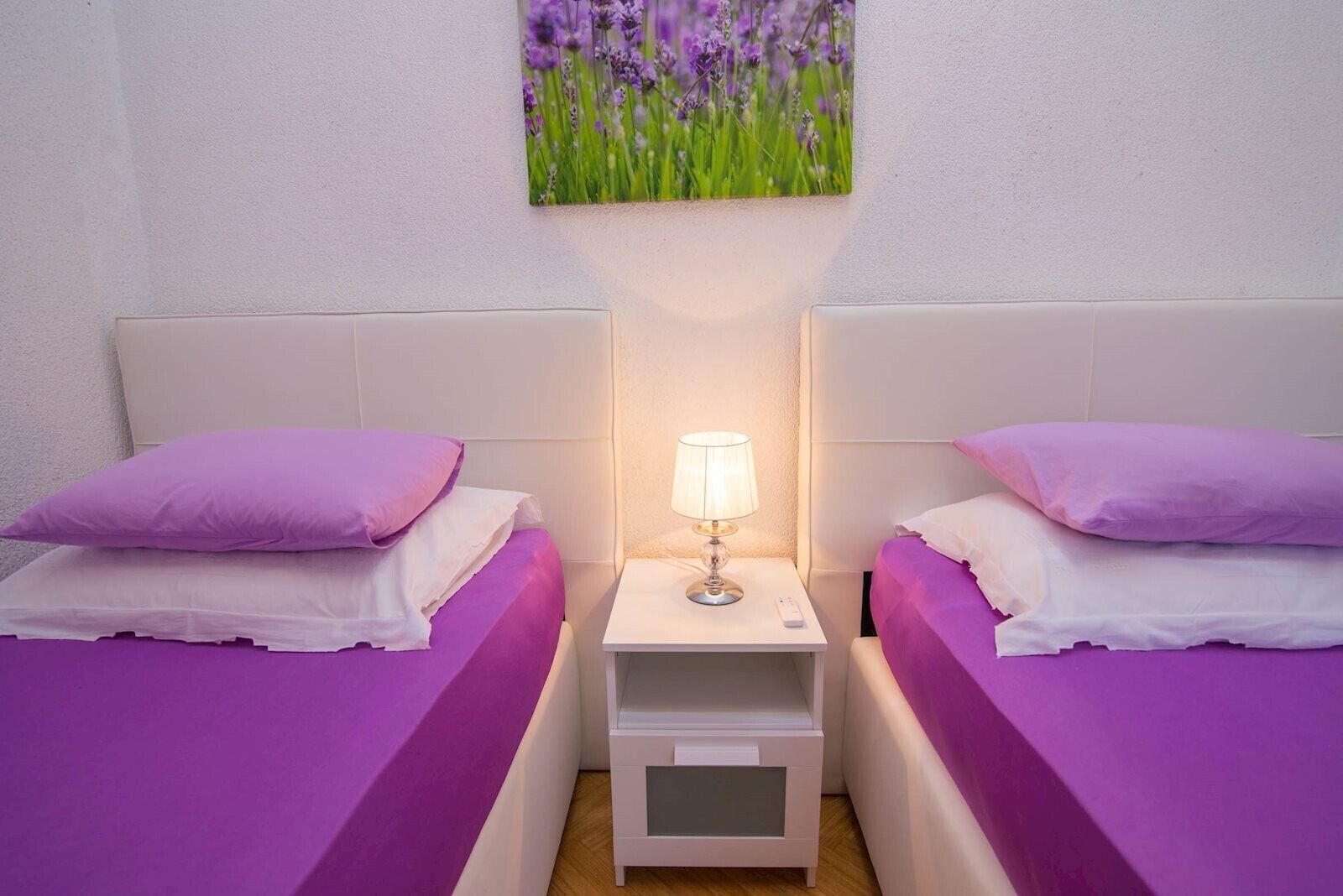 A room with two beds, each with a purple bedspread and matching pillows. There is a nightstand with a lamp between the beds, and a painting of flowers on the wall above the nightstand.