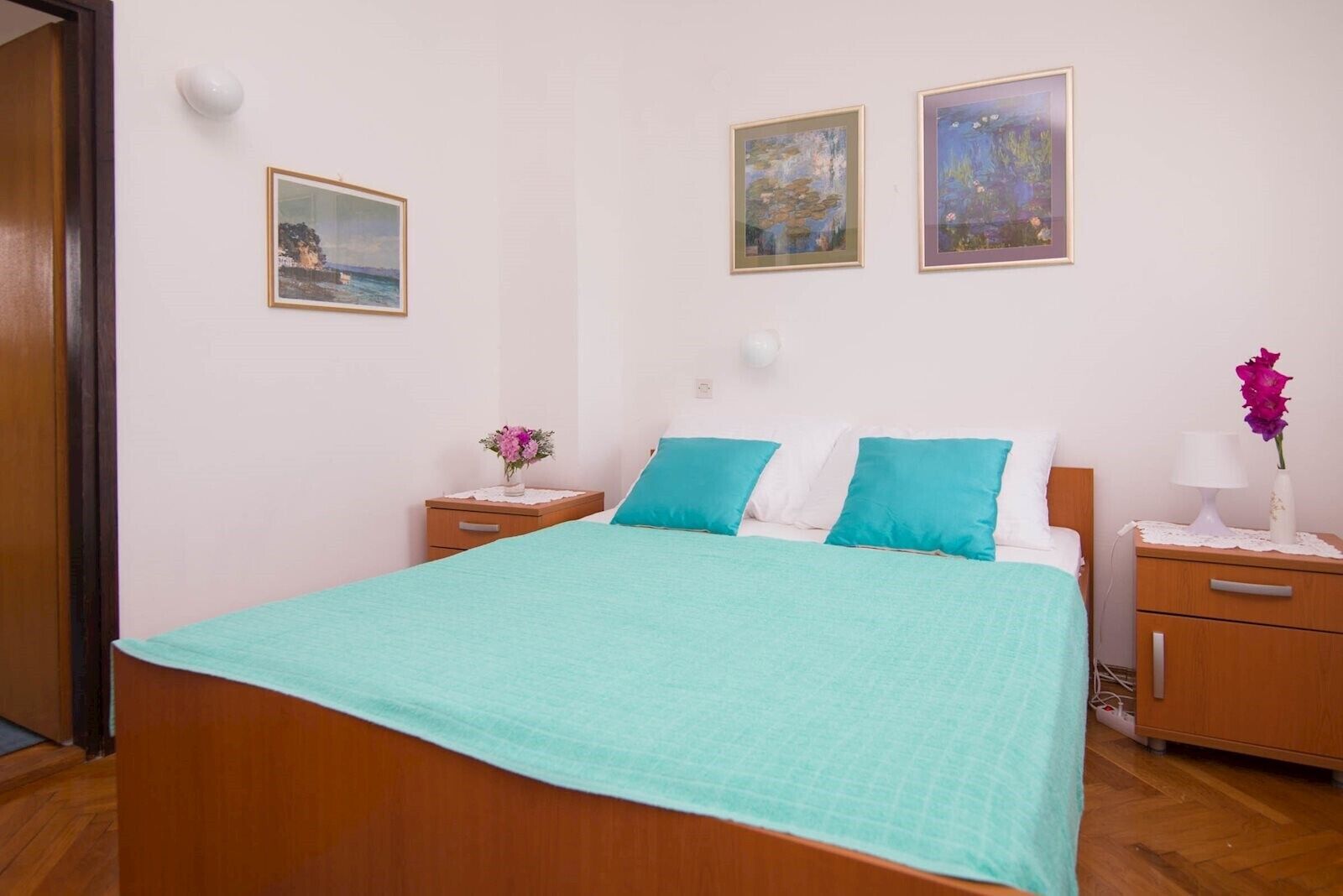 Een slaapkamer met een bed, twee nachtkastjes, een houten deur en drie ingelijste kunstwerken aan de muur.
