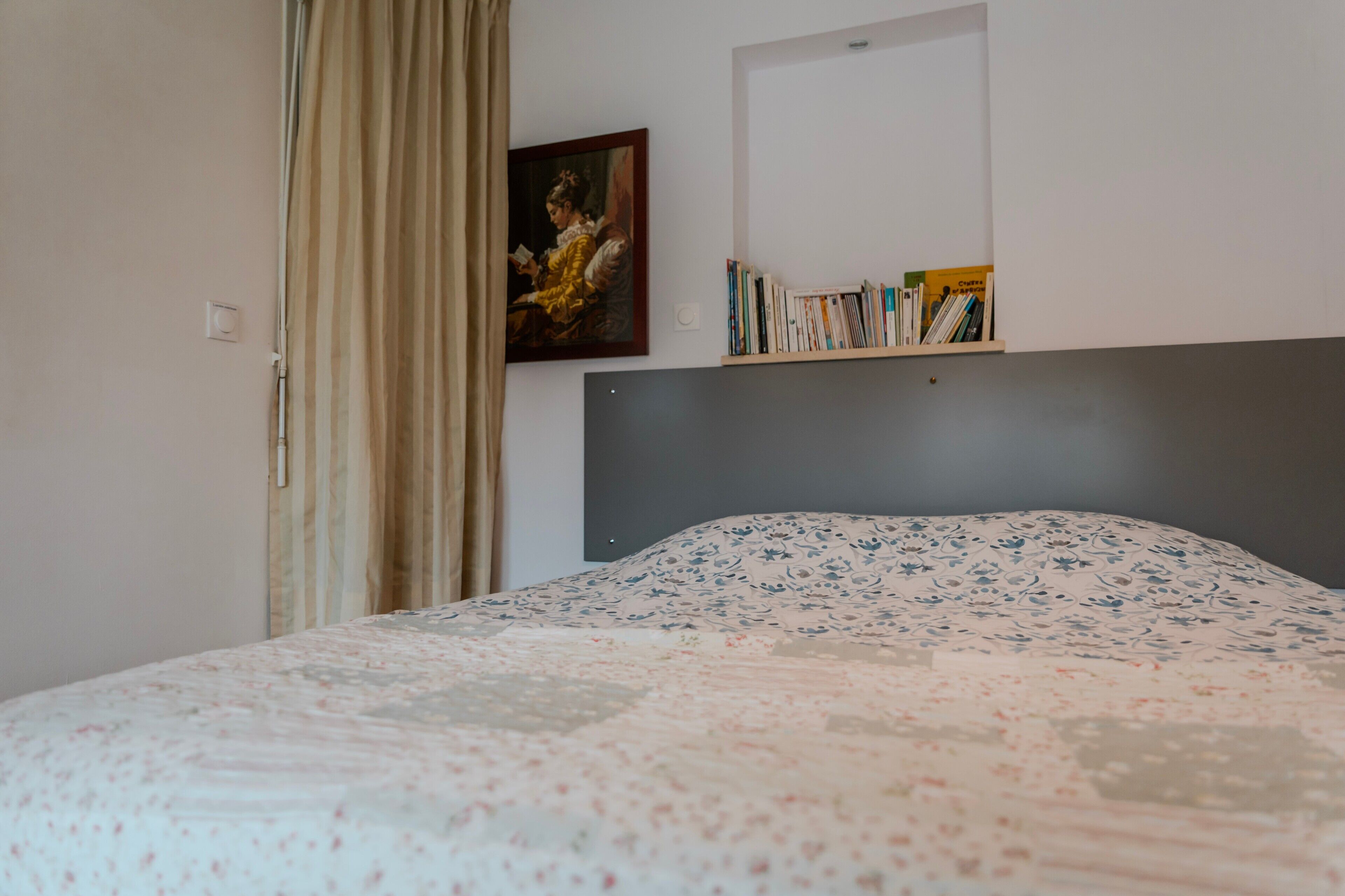 A bedroom with a bed, a painting, a bookshelf, and a window with curtains.
