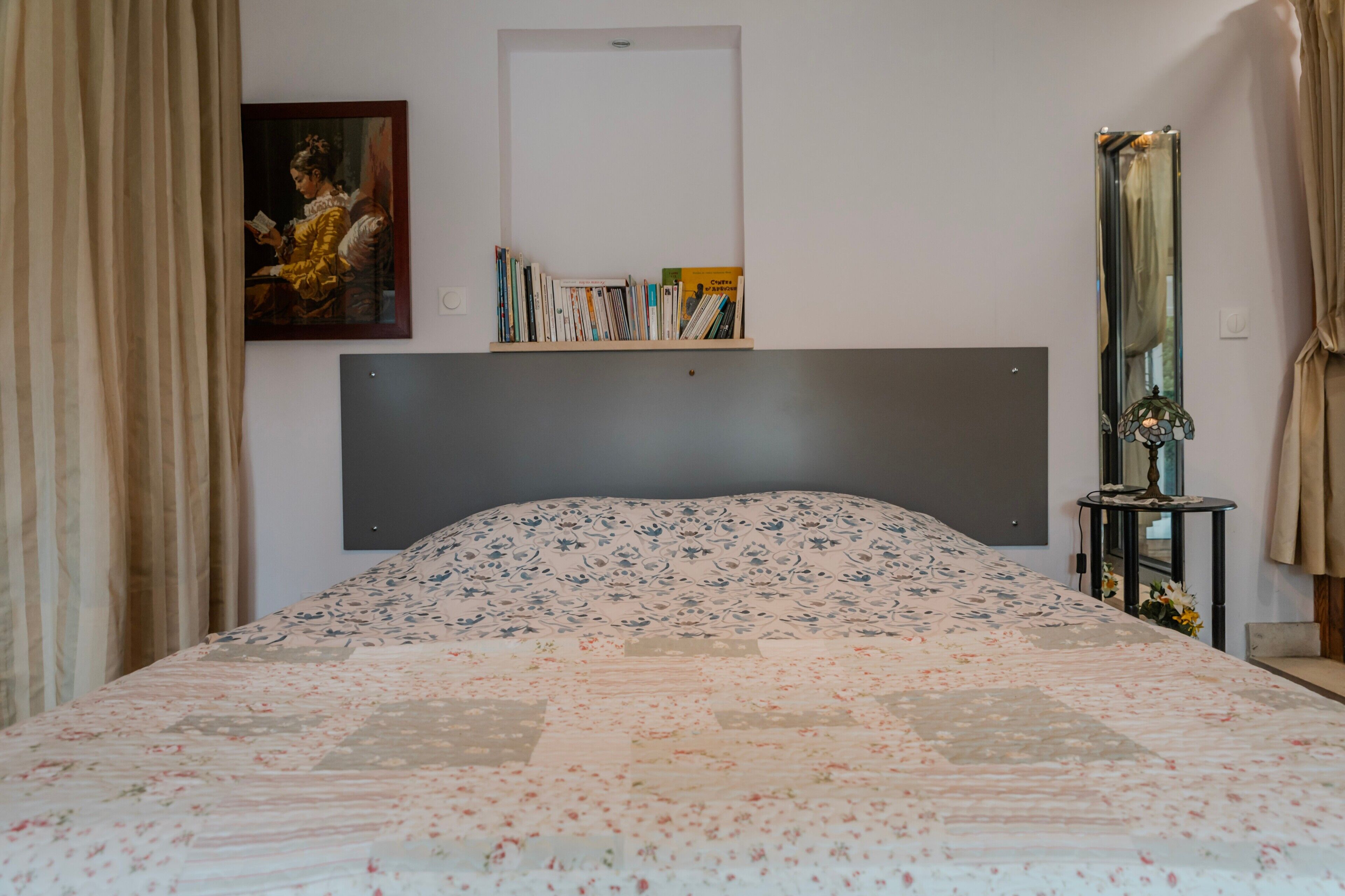 A bedroom with a bed, a bookshelf, a painting, a mirror, and a side table.