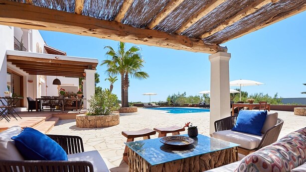 Een zwembadterras met buitenmeubilair, een houten pergola en uitzicht op het zwembad en palmbomen.