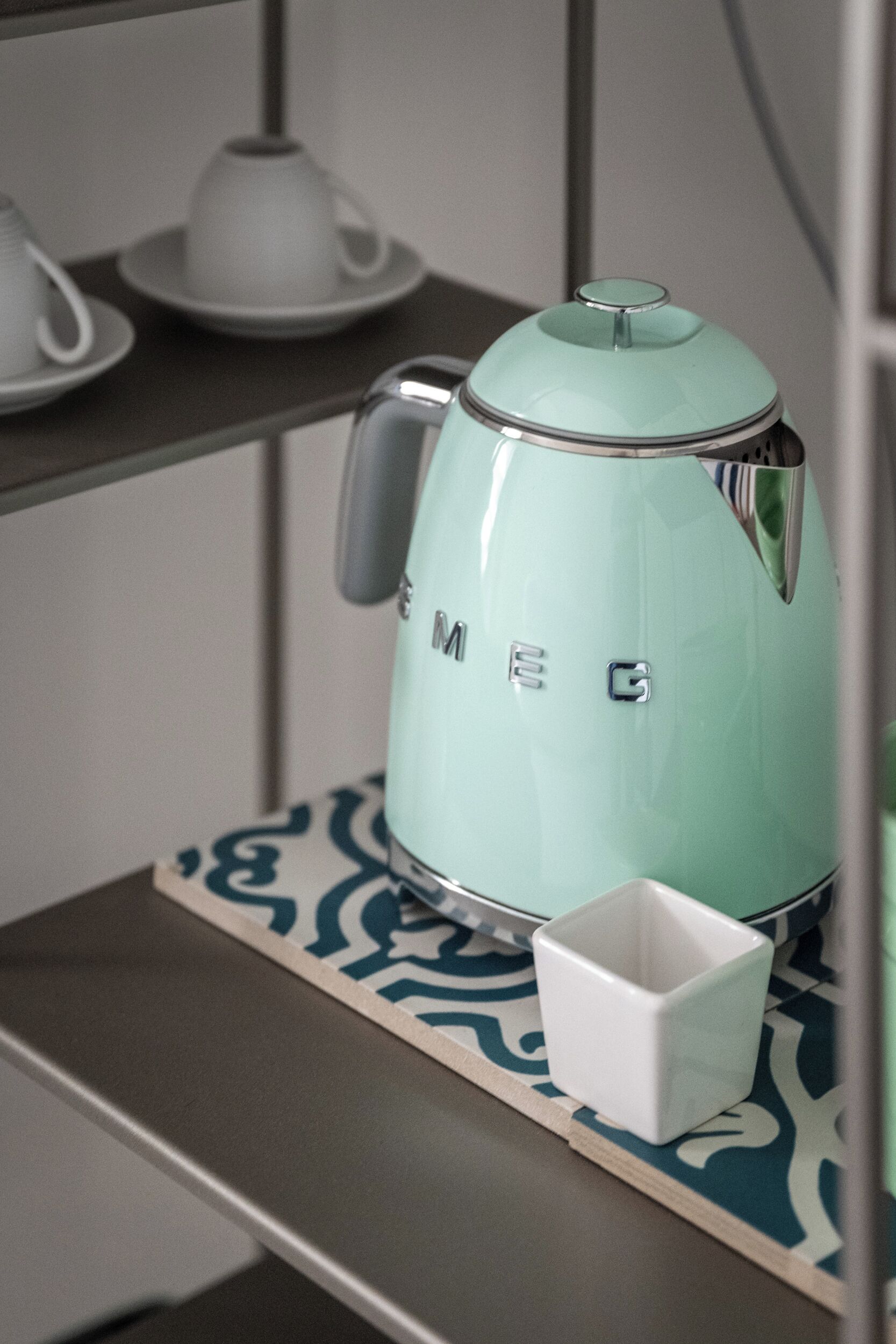 Una màquina de cafè de color blau verd amb el nom de marca “MEG” sobre una prestatgeria.