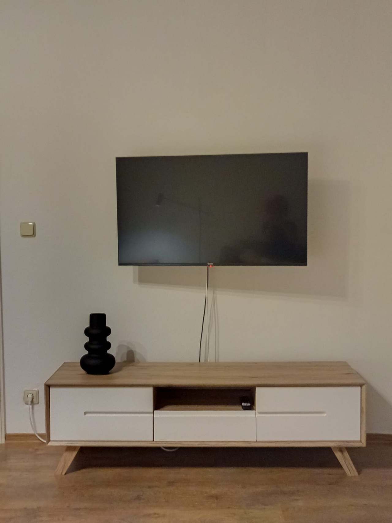 Televisão de tela plana fixada na parede acima de um móvel de madeira com duas gavetas.