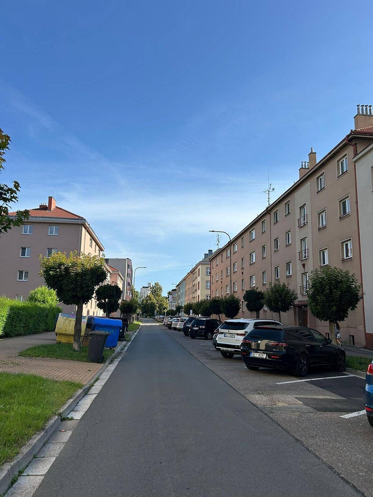 A residential street lined with multi-story buildings, parked cars, and small trees along the sidewalk.