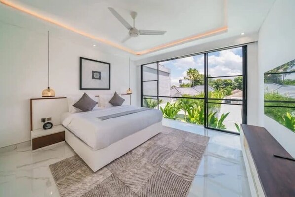 A modern bedroom with a large bed, a ceiling fan, and a view of greenery outside.