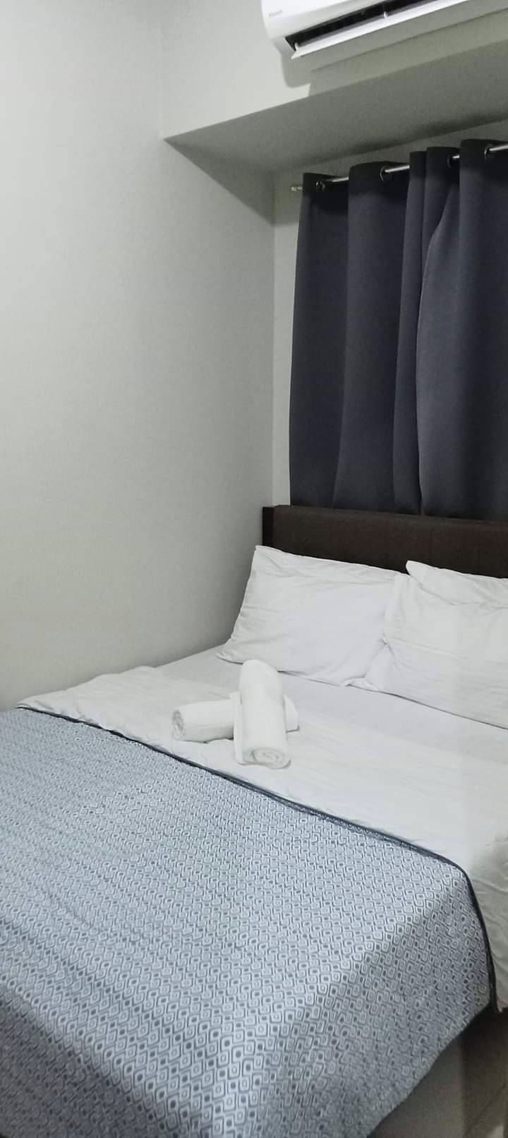 A hotel room with a bed, white linens, a grey patterned bedspread, a dark curtain, and an air conditioning unit above the bed.