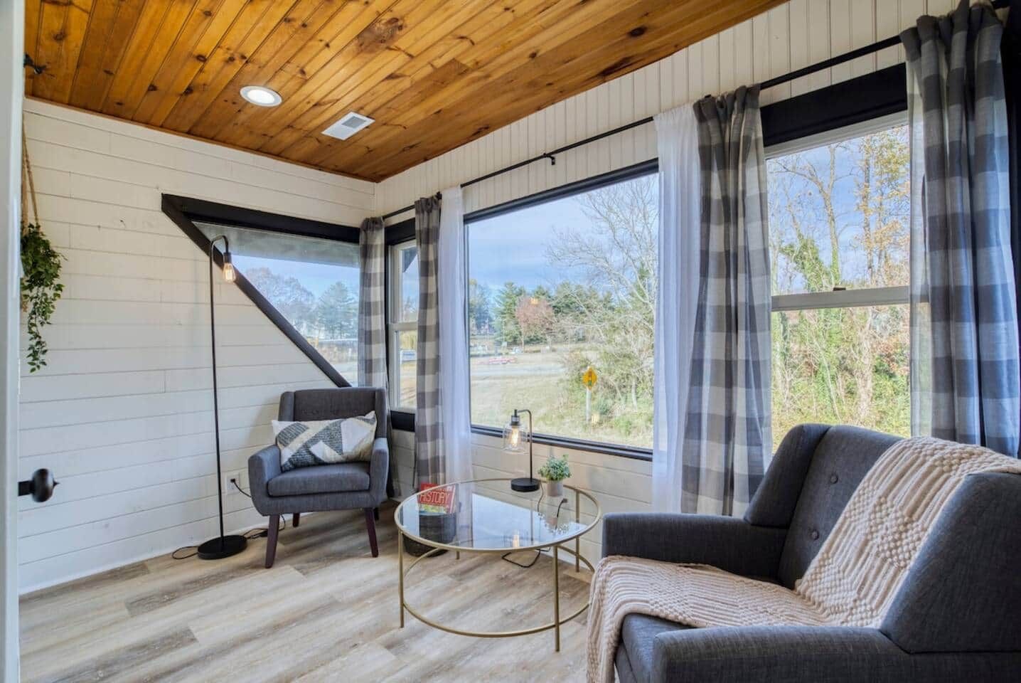Une pièce avec un plafond en bois, de grandes fenêtres avec des rideaux, un canapé, un fauteuil et une petite table.